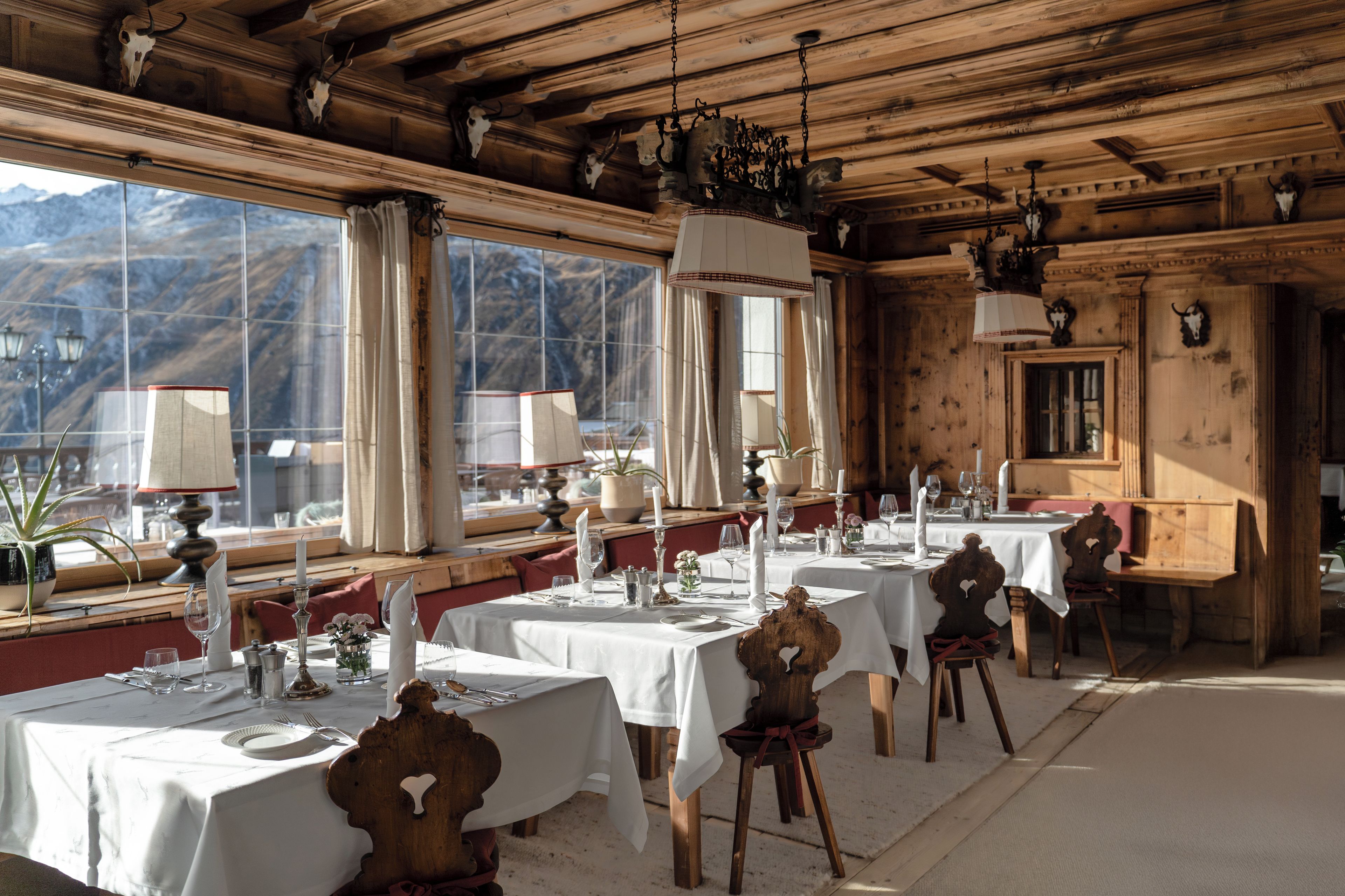 Uma sala de jantar com várias mesas preparadas para uma refeição, grandes janelas com vista para as montanhas e vigas de madeira no teto.