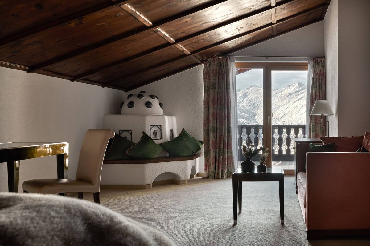 Un cuarto acogedor con techo de madera, un sofá, una silla, una mesa y vista a montañas a través de un ventanal grande.