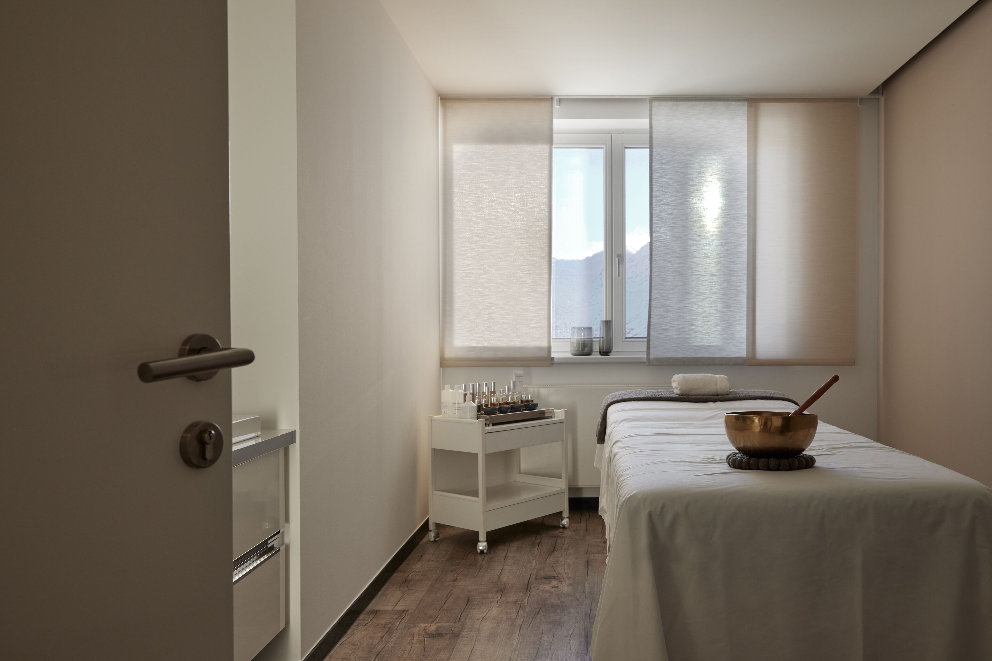 Una camera da letto con un letto, un comodino con una ciotola e un cucchiaio, una finestra vista montagne e una porta con maniglia.