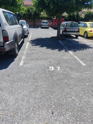 Ein Parkplatz mit mehreren Autos, darunter ein weißer SUV, ein silbernes Auto und ein gelbes Auto. Im Hintergrund sind Bäume und ein Gebäude mit einem Ziegeldach zu sehen.