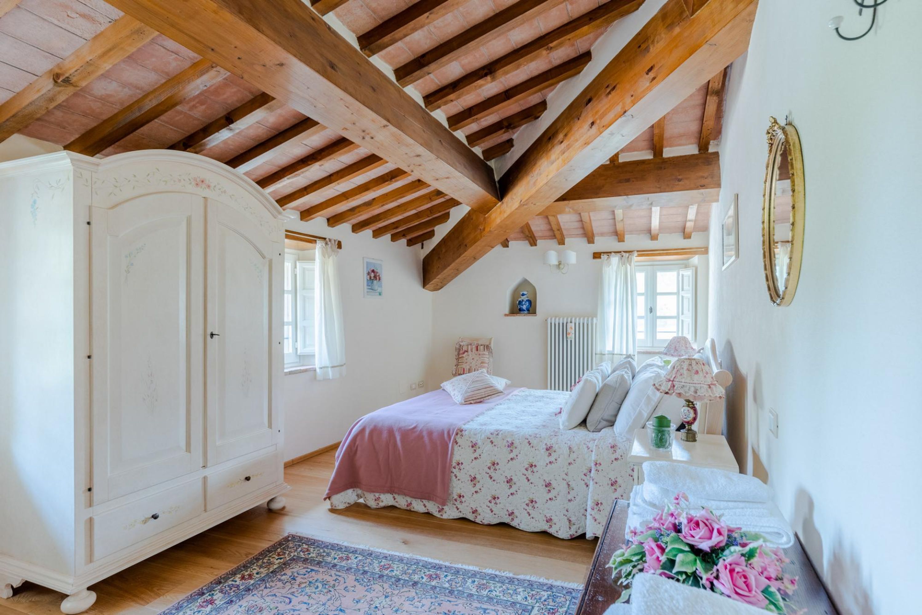 A bedroom with a wooden ceiling, a bed with floral bedding, a white wardrobe, and a small table with a lamp.