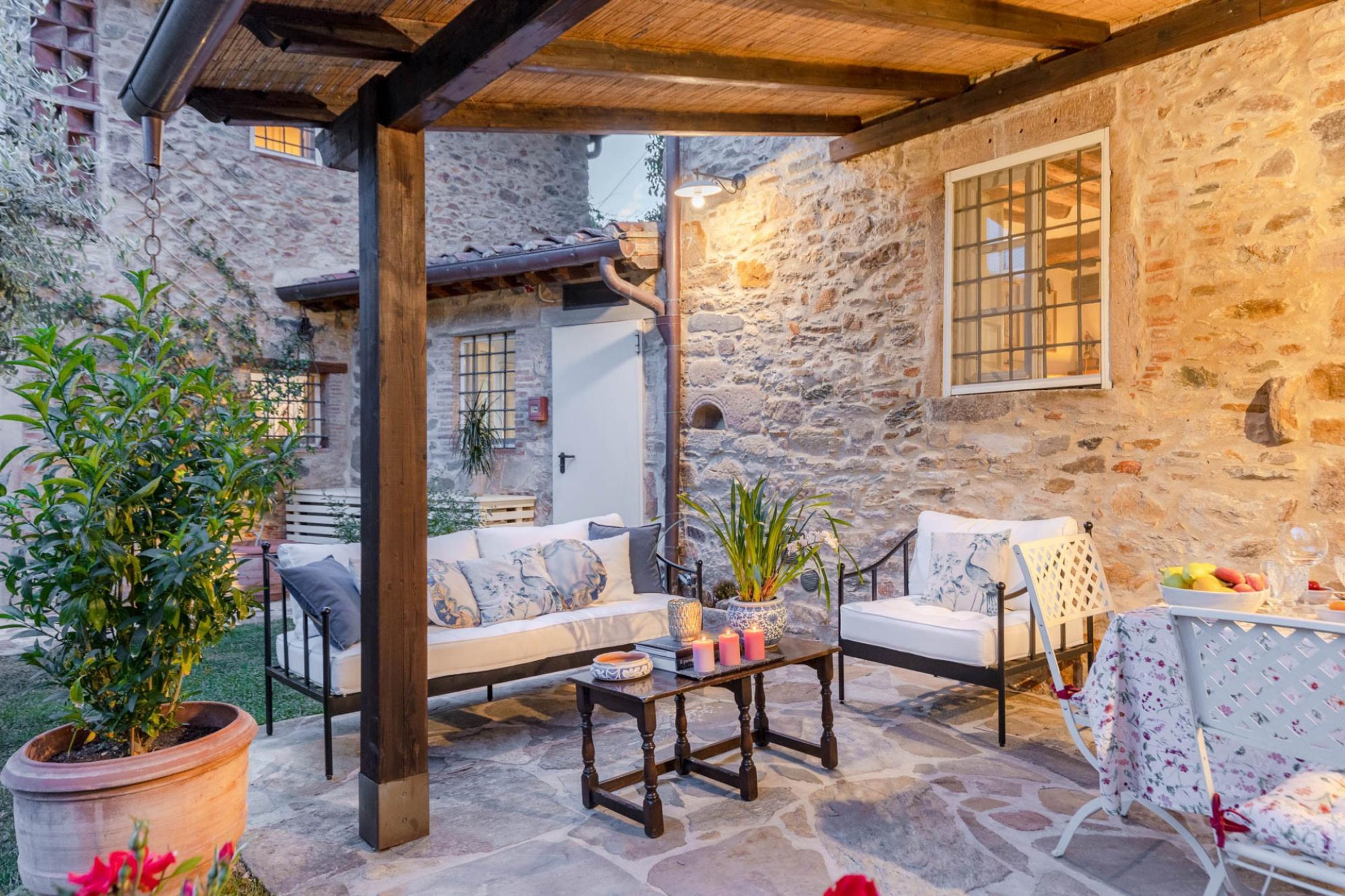 A stone-walled patio with a wooden pergola, outdoor seating, and potted plants.