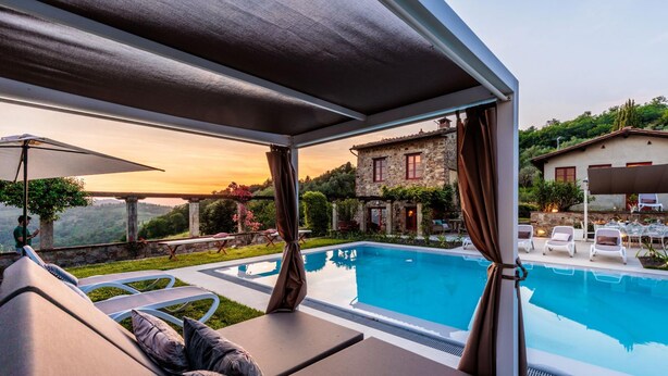 Ein Poolbereich mit Liegestühlen, einer überdachten Sitzgelegenheit und Blick auf ein Steinhaus sowie üppiges Grün.