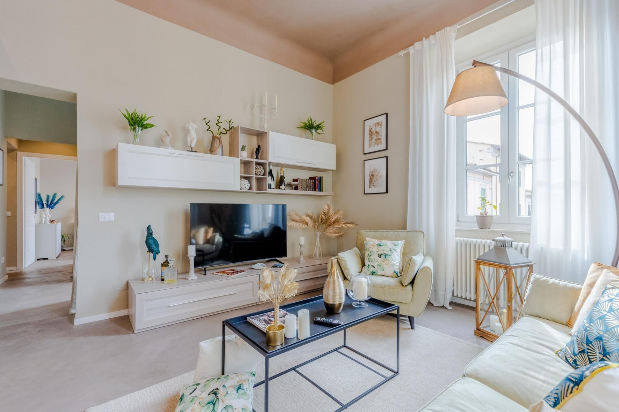 A modern living room with a television, white sofas, a coffee table, and a large window with curtains.