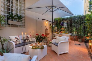 Ein Patio mit weißen Korbmöbeln, einem großen Sonnenschirm und einem Esstisch, gedeckt mit Kerzen und Blumen.