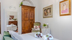Une chambre à coucher comprenant un lit, une armoire en bois, une chaise, une petite table avec un vase, et deux œuvres d’art encadrées accrochées au mur.