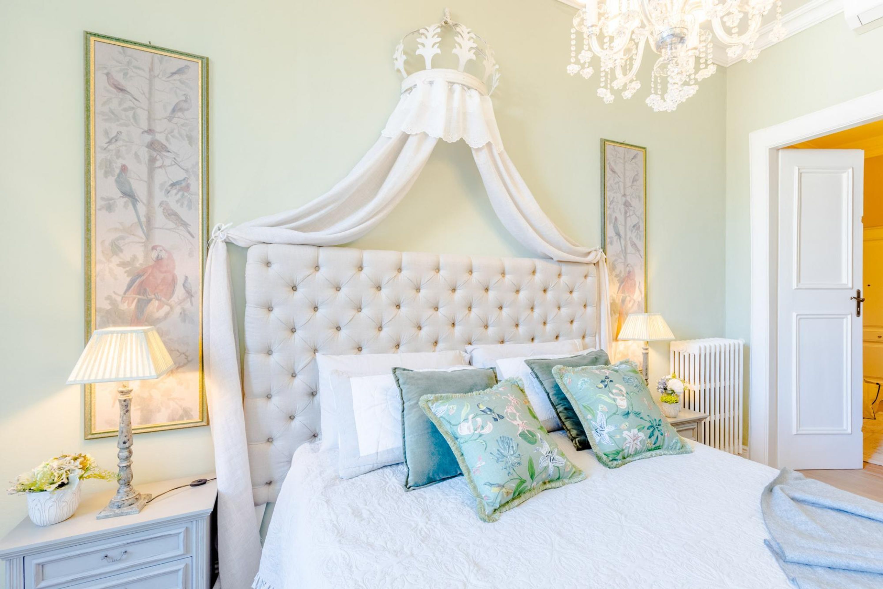 Un dormitorio con cabecera capitonada, cama con dosel, almohadas estampadas, una mesita de noche con lámpara y un cuadro en la pared.
