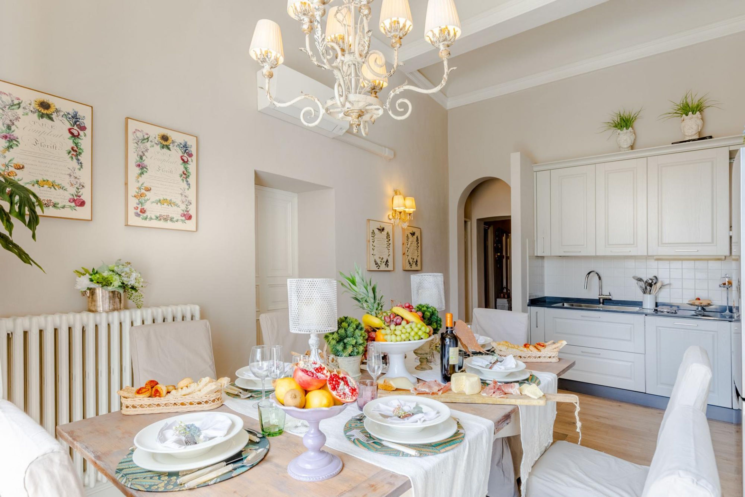 A well-lit dining area with a chandelier, white cabinets, and a fruit centerpiece on the table.