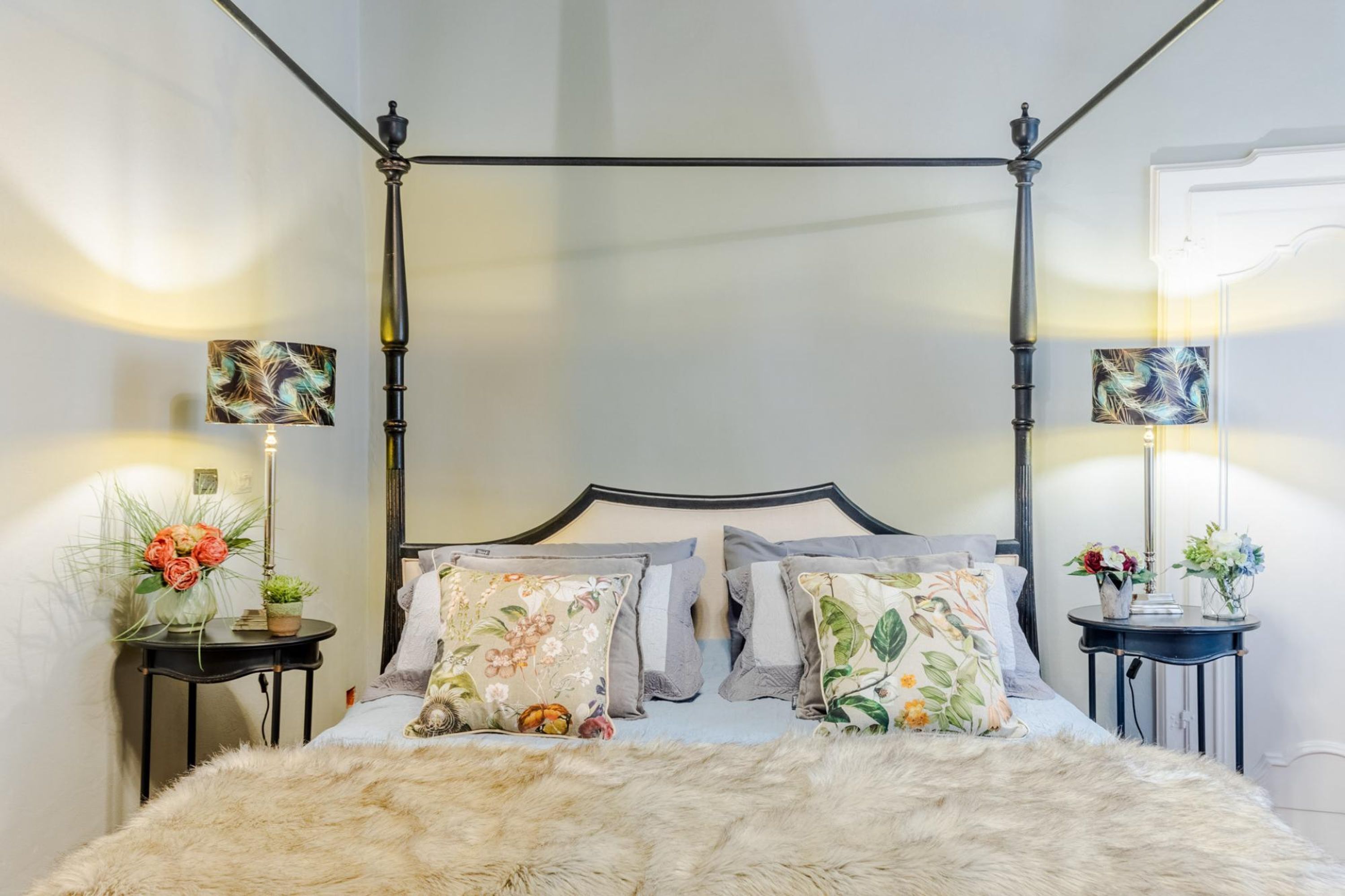 Uma cama com dossel adornada com almofadas florais, um edredom branco e fofo, e duas mesas de cabeceira com abajures e arranjos florais.