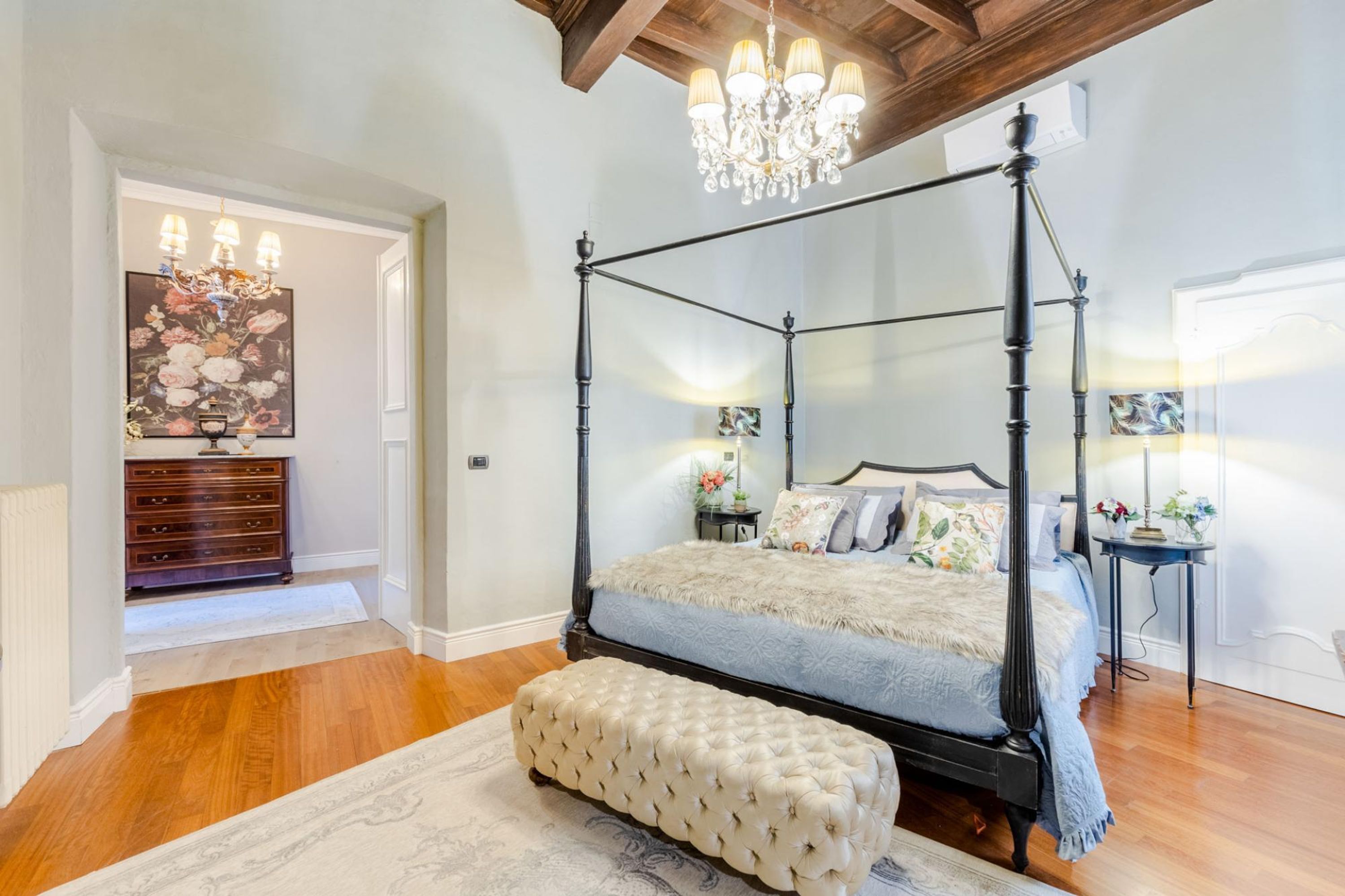 Uma cama de dossel com um banquinho estofado num quarto com soalho em madeira e um candelabro.