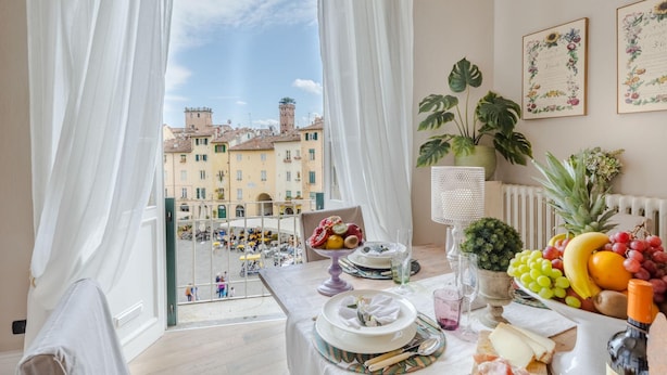 Un'area pranzo con un tavolo apparecchiato per due, vista sulla città attraverso ampie finestre e un balcone con persone.