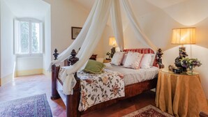 Un dormitorio con cama a dosel, dos lámparas de noche, una mesita de noche con lámpara y una ventana con cortinas.