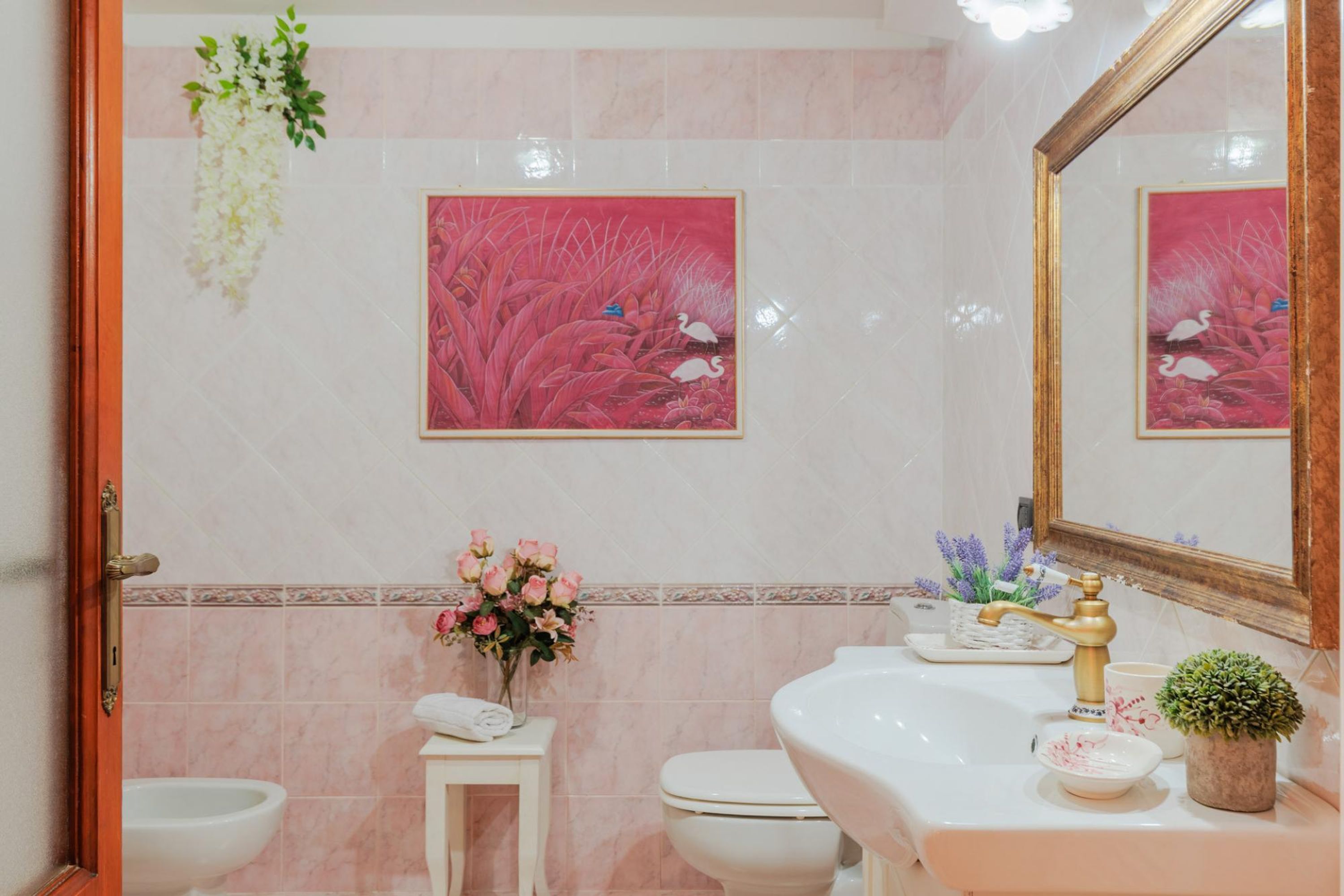 A bathroom with a white sink, a gold faucet, a mirror, and a toilet. There are pink and white flowers on a small table.
