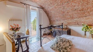 A bedroom with a brick ceiling, a bed with a white bedspread, a wooden chair, a table with a vase of flowers, and a window with a curtain. - Casa La Guardia a 3 Bedrooms Panoramic Apartment (Lucca)