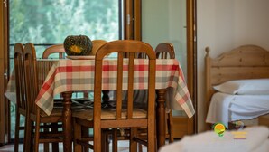 Un comedor con mantel a cuadros, sillas de madera y una calabaza sobre la mesa. Se ve una cama con ropa blanca de fondo.