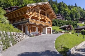 A wooden chalet with a balcony, set in a mountainous area with a stone wall and a paved path leading to it.