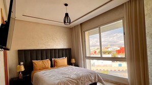 Ein modernes Schlafzimmer mit einem großen Bett, einem Flachbildfernseher, einem Nachttisch und Blick auf die Stadt durch ein großes Fenster.
