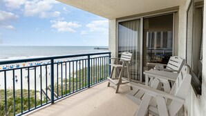 A balcony with a view of the beach and ocean, featuring two white chairs and a table.