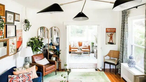 Un salon chaleureux avec un canapé bleu, un fauteuil marron et un tapis vert. Il y a trois miroirs au mur, un ventilateur de plafond noir et une fenêtre avec des rideaux.