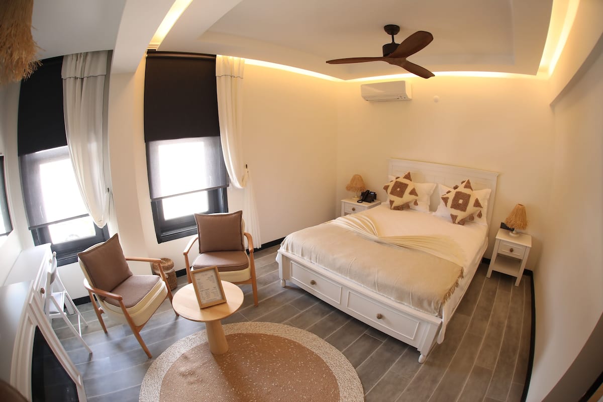 A bedroom with a bed, two chairs, a small table, a ceiling fan, and a window with curtains.