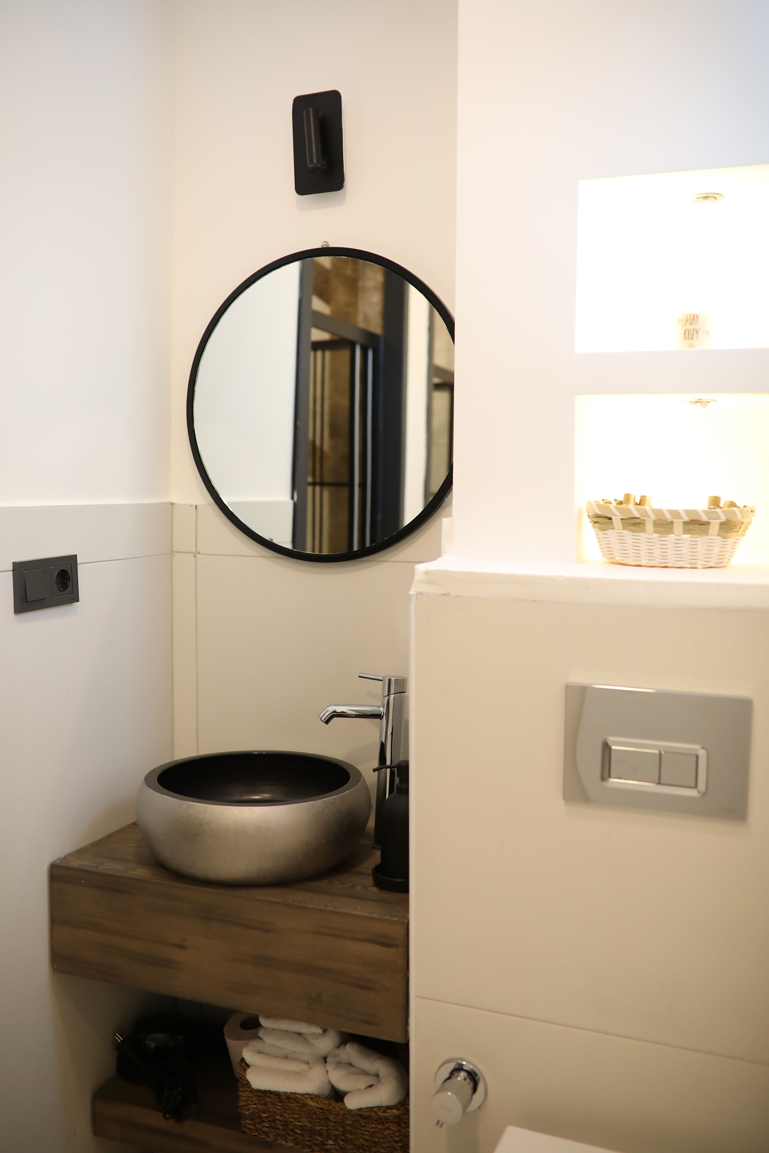Un baño con un espejo redondo, un lavabo con un recipiente, una cesta de toallas y una lámpara.