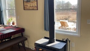 Ein Zimmer mit Holzofen, einem Fenster mit Blick ins Freie, einem Bild an der Wand und einem Tisch mit einer Bank.