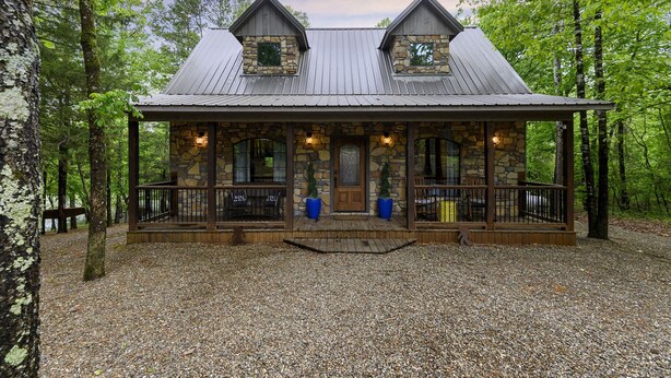 A rustic cabin with a stone facade, wooden porch, and metal roof, surrounded by trees.