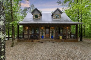 A rustic cabin with a stone facade, wooden porch, and metal roof, surrounded by trees. - Private Pond • Hot Tub • Paddle Boat • 2 Kings (Broken Bow)