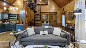 A cozy log cabin interior with a gray sofa, a wooden staircase, a kitchen area, and a dining table.
