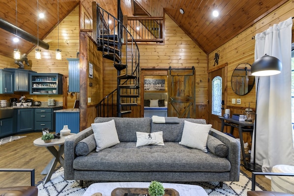 L’intérieur d  ’un chalet confortable, avec un canapé gris, un escalier en bois, un coin cuisine et une table à manger.