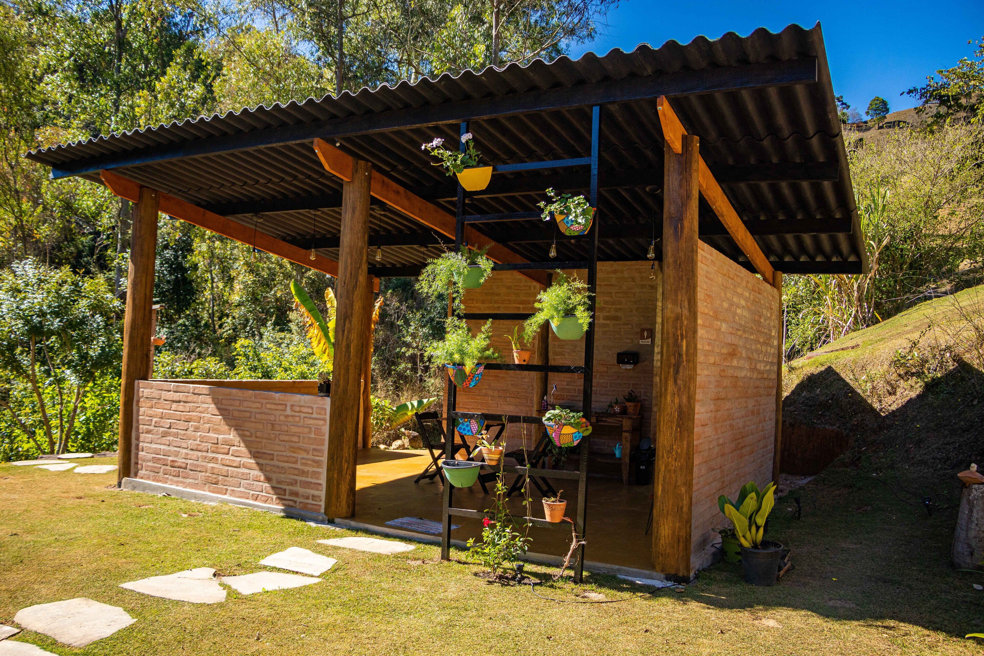 Uma pequena estrutura ao ar livre com telhado de metal corrugado e paredes de tijolo, rodeada por um jardim com plantas em vasos e um caminho de pedra.