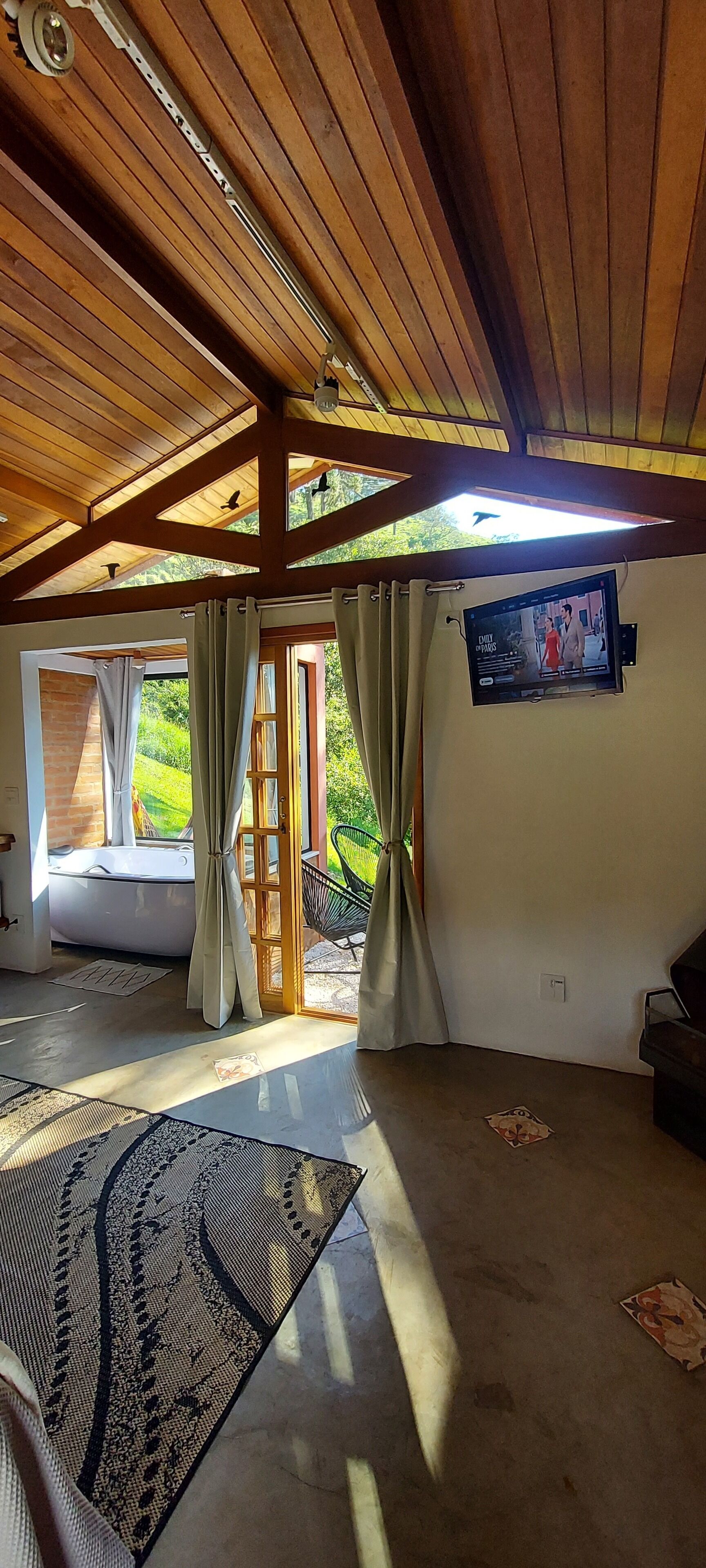 A room with a wooden ceiling, a large window, a flat-screen TV, a bathtub, and a patterned rug.