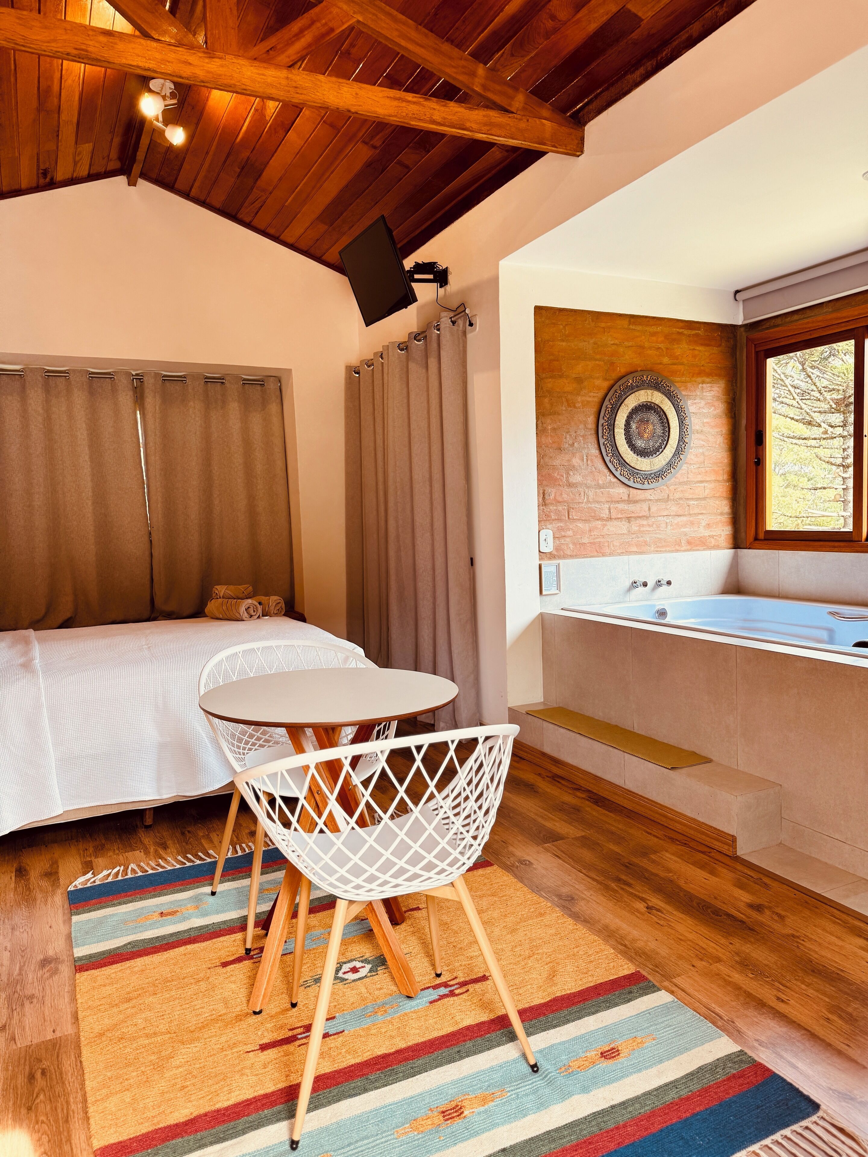 Une pièce confortable avec un lit, une petite table ronde, une chaise blanche, un espace douche et une fenêtre donnant sur des arbres.