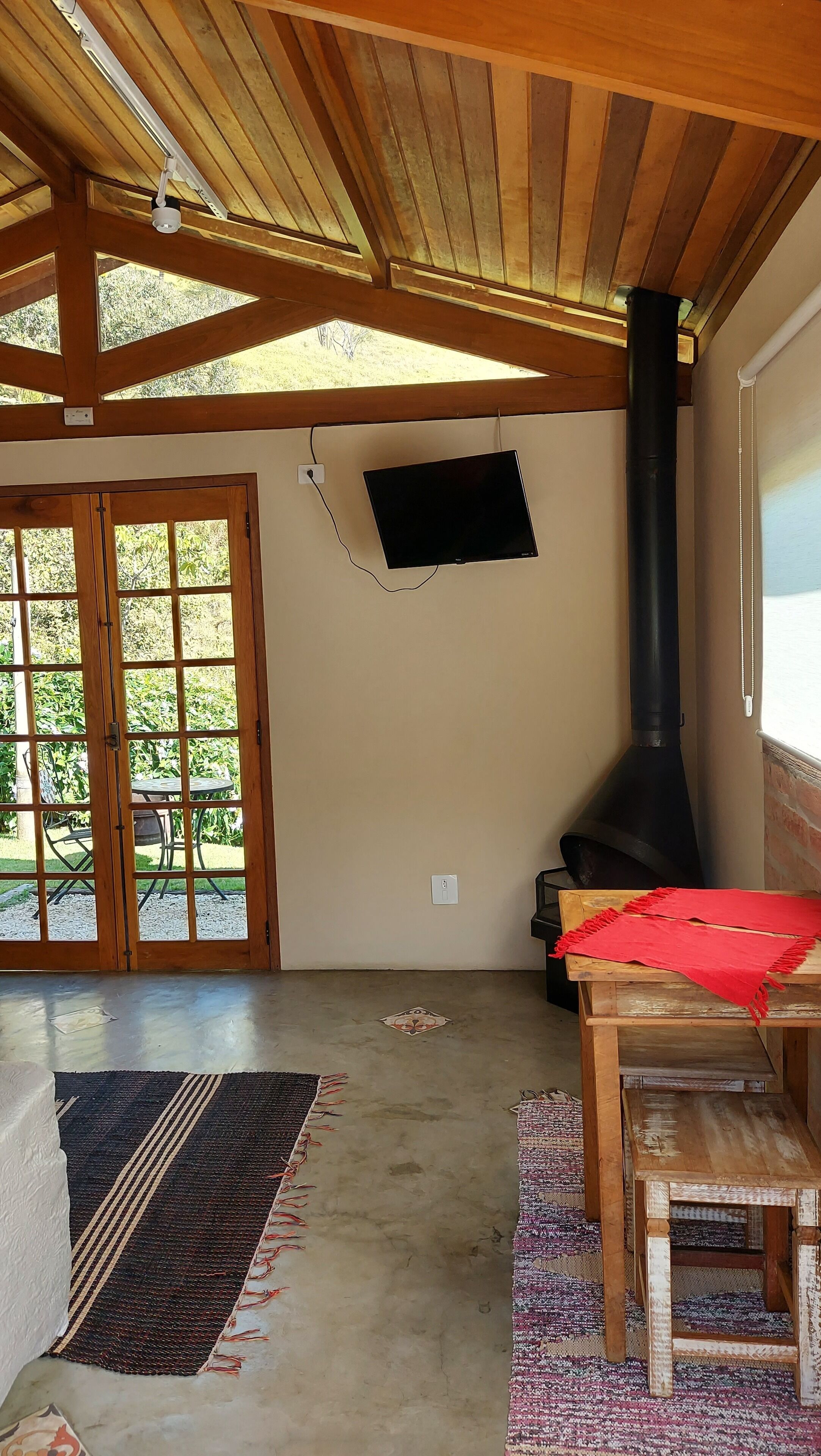 A spacious room with a high ceiling, a black stove, a TV mounted on the wall, and a large wooden door leading to an outdoor area.