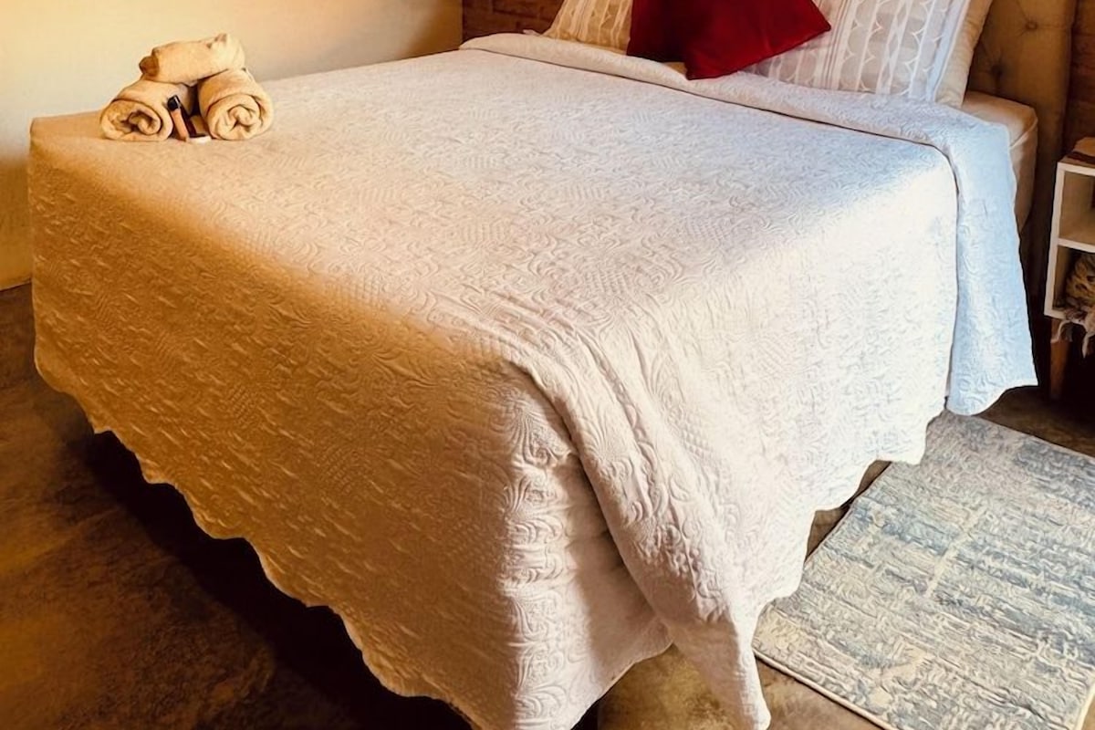 A single bed with a white textured bedspread, a red pillow, and a headboard against a brick wall.