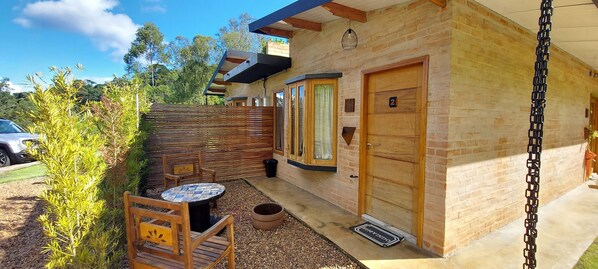 Un pequeño edificio de ladrillo con una puerta de madera marcada con el número '2', un pequeño patio con una mesa y sillas, y una valla de madera.