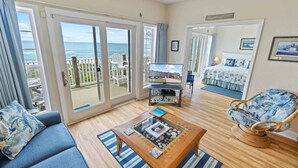 Una stanza con un'ampia porta-finestra che si apre su un balcone con vista sull'oceano. C'è un letto con una coperta a tema spiaggia in blu e bianco, una televisione su un mobile e una sedia di vimini con cuscino.