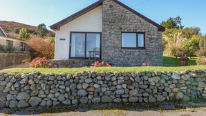 A two-story stone house with a sloped roof, a stone wall in front, and a grassy yard. - Hefo'r Grug (Pwllheli)