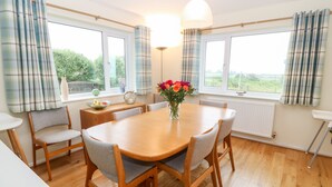 A dining area with a wooden table and chairs, a vase with flowers, and large windows with curtains. - Hefo'r Grug (Pwllheli)
