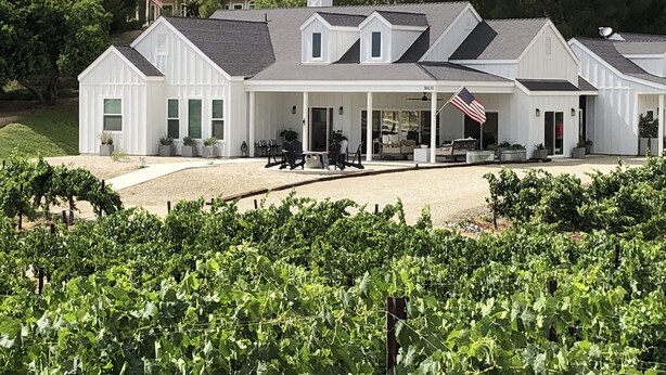 A large white building with a covered patio area and a vineyard in the foreground. - Lux Vineyard Escapes-Ultimate Modern Farm House (Temecula)