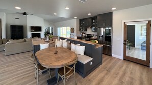 A modern kitchen with dark cabinets, a stainless steel refrigerator, and a dining area with a wooden table and chairs. - Lux Vineyard Escapes-Ultimate Modern Farm House (Temecula)