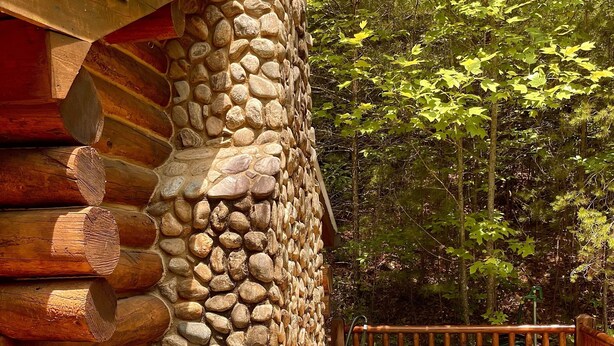 Une cabane en rondins avec une façade en pierre, un jacuzzi et un barbecue sur une terrasse en bois.