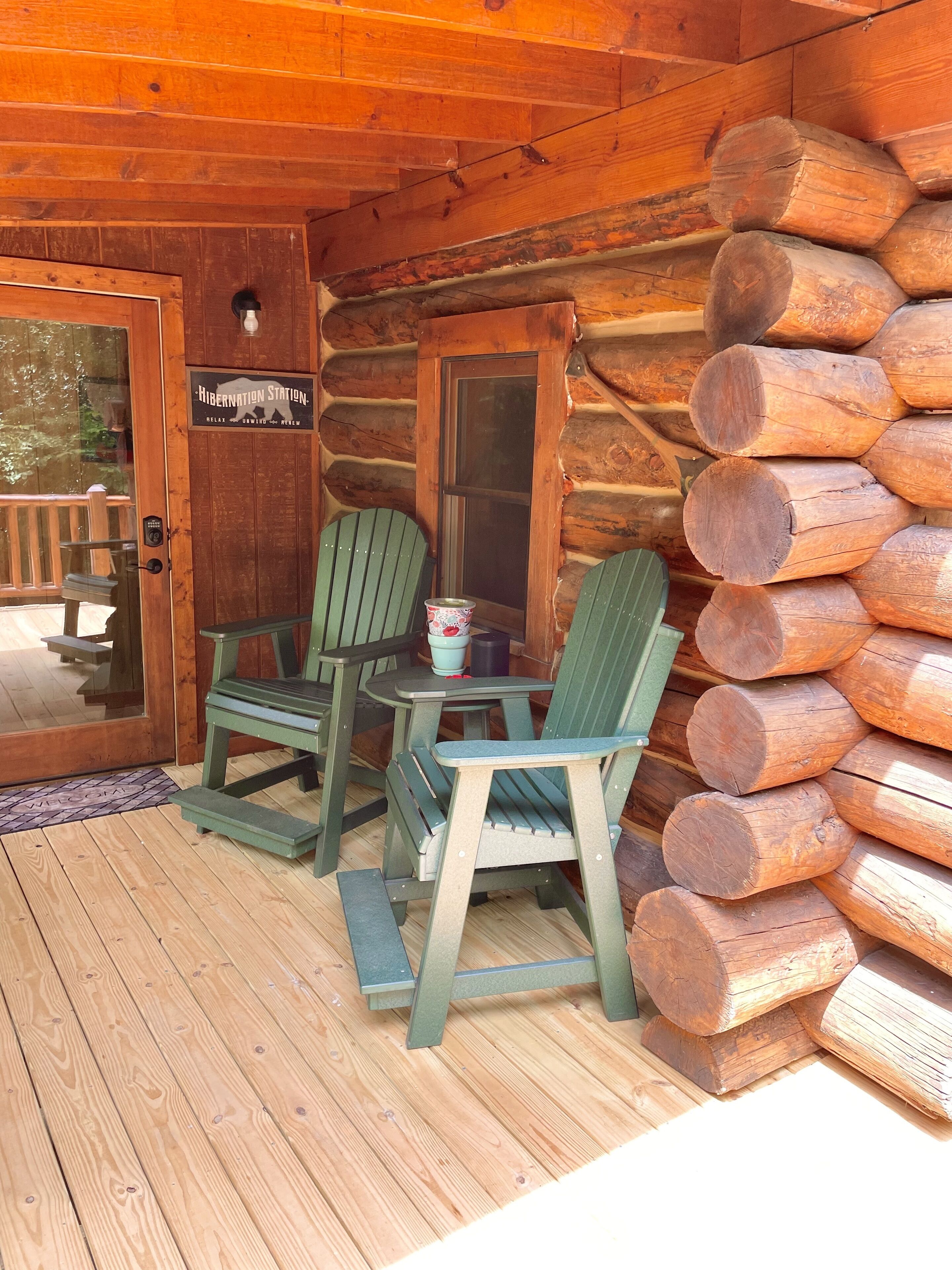 A wooden cabin with a porch featuring two green Adirondack chairs and a small table. A sign on the door reads "Hibernation Station."