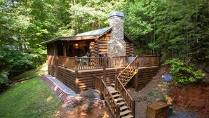 Une cabane en rondins avec une cheminée en pierre, une terrasse en bois et un escalier menant à l’entrée.