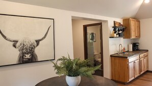 Une cuisine moderne avec des meubles en bois, un évier et une machine à café. Une plante en pot est posée sur une table ronde, et un tableau représentant un taureau est accroché au mur.