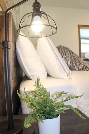 A bed with white bedding, a potted fern, and a floor lamp with a light bulb. - Horse Creek Resort - Cabin 12 (Rapid City)