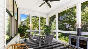 Une terrasse couverte avec une table et des chaises, une cheminée et de grandes fenêtres donnant sur un espace verdoyant.