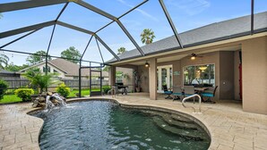 A swimming pool with a waterfall feature, surrounded by a covered patio area with outdoor furniture and a view of the backyard.