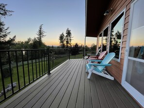 Une terrasse en bois avec une rambarde, deux fauteuils Adirondack et une vue sur les arbres et le coucher du soleil.