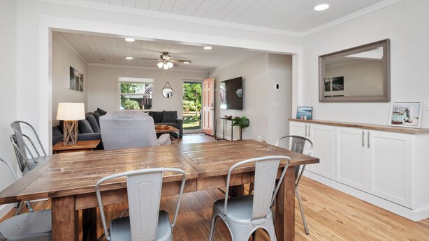 A dining area with a wooden table and metal chairs, a kitchen with white cabinets, and a ceiling fan. - Beach Please: Modern Coastal Bungalow Near Waterway and Marshwalk (Oak Island)